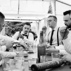 Panowie w eleganckich koszulach i szelkach świętują kawalerski pomysły przy stole z cygarami i drinkami.