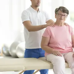 Starsza pani na rehabilitacji. Fizjoterapeuta wspiera ją podczas ćwiczeń w sanatorium ortopedycznym NFZ.