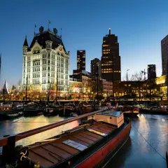 Wieczorem w porcie, gdzie jest Rotterdam. Widać zabytkowy budynek, nowoczesne wieżowce i zacumowane łodzie.