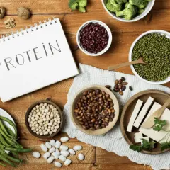 Notatnik z napisem "PROTEIN" otoczony miseczkami z fasolą, ciecierzycą, soczewicą, orzechami, brokułami, fasolką szparagową i tofu.
