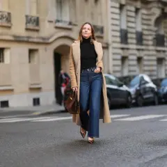Une femme porte un jean flare bleu, idéal pour allonger la silhouette et sublimer toutes les morphologies. Elle est habillée d'un long manteau camel et d'un col roulé noir.