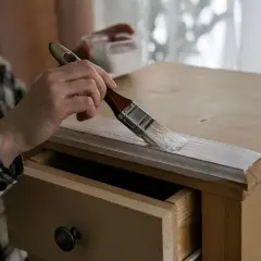 Persona con camisa a cuadros pinta muebles de madera con brocha blanca.