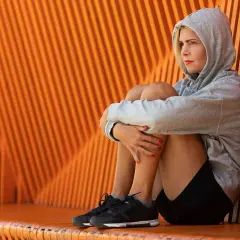 En kvinna i grå hoodie sitter hopkrupen mot en orange vägg, en bild av mental styrka trots en stunds eftertanke.