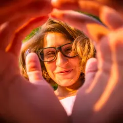 Ragazza con occhiali, sorride attraverso le mani che creano una cornice. Un'esperienza emozionale correttiva che illumina il volto.