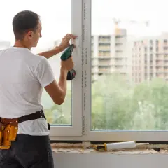 Montaż okna trwa, fachowiec używa wkrętarki. W tle widać budowę.