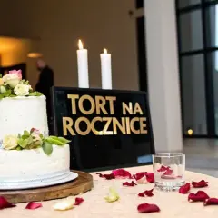 Elegancki tort na rocznicę ślubu, ozdobiony kwiatami, stoi obok tablicy z napisem "TORT NA ROCZNICĘ" i płonących świec.