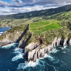 Zielone pola i skaliste klify na wyspach należących do Portugalii, z błękitnym oceanem i chmurami na niebie.