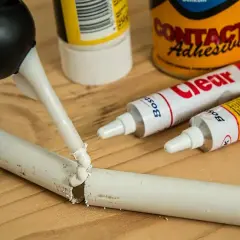 Pistolet z klejem aplikuje masę na pękniętą białą rurkę. Obok leżą tubki kleju, pokazujące, czym skleić plastik.