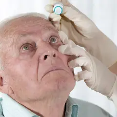 Starszy mężczyzna z zielonymi oczami otrzymuje krople do oczu. Rękawiczki medyczne, **yellox** na opakowaniu.
