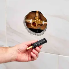 Hand holding a shower valve cartridge next to an open wall cavity revealing the shower valve body.