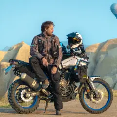 Mężczyzna w kurtce Revit Sand 4 siedzi na motocyklu terenowym na tle skalistych formacji i balonu na gorące powietrze.