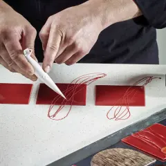 Mains d'un pâtissier créant un décor royal chocolat sur des carrés rouges brillants.