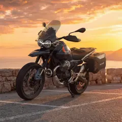 Moto Guzzi V85 TT lista para la aventura al atardecer, con su diseño robusto y maletas laterales, frente a un mar sereno y montañas doradas.