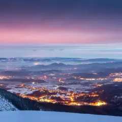 Zachód słońca nad Karkonoszami. Rozświetlone miasto w dolinie, ośnieżone zbocza i zimowe szlaki tworzą magiczny krajobraz.
