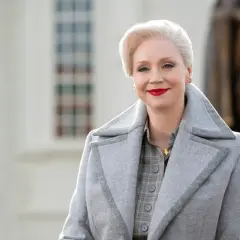 Dyrektorka z Wednesday, Larissa Weems, w eleganckim szarym płaszczu i z czerwoną szminką.