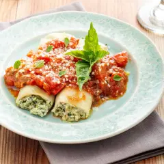 Canelones de espinacas cubiertos con salsa de tomate y queso rallado, adornados con hojas de albahaca fresca.