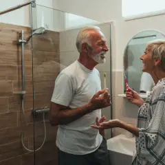 Ein älteres Paar putzt sich die Zähne. Sie lachen und scheinen sich über den Umbau des Badezimmers zu freuen, der ihnen den zuschuss badumbau pflegestufe 1 ermöglicht hat.