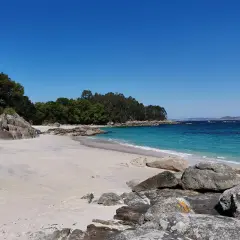 Arena blanca, aguas turquesas y rocas salpican la **playa de Castiñeiras**, un rincón idílico bajo un cielo azul intenso.
