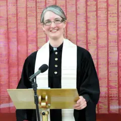 Eine Pfarrerin mit Brille und grauem Haar steht lächelnd hinter einem goldenen Pult. Sie trägt eine schwarze Robe und ein weißes Stola. In dir steckt ein guter Christ.