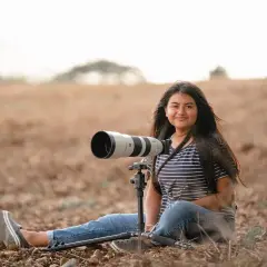 Młoda kobieta z długimi włosami, w pasiastym T-shircie i jeansach, siedzi na ziemi w polu z profesjonalnym aparatem na statywie. To jej fotografia lifestylowa.