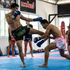 Two Muay Thai fighters spar, one delivering a powerful kick. A coach observes, ready to check hook technique.