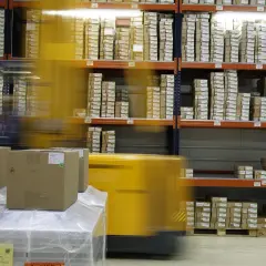 Wózek widłowy porusza się w magazynie, gdzie na regałach piętrzy się pole odkładcze z paczkami.