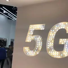 Wielkie, podświetlane litery "5G" na ścianie, symbolizujące przyszłość technologii. W tle widać ludzi i stoiska targowe.