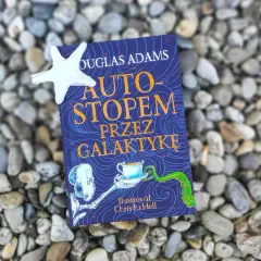 Okładka książki "Autostopem przez galaktykę" Douglasa Adamsa, z ilustracją Chrisa Riddella, leży na kamieniach.