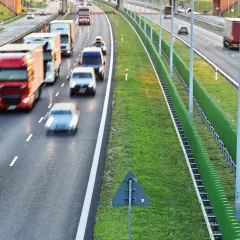 Autostrada z ruchem samochodowym i ciężarowym, zielone ekrany akustyczne i znak drogowy.