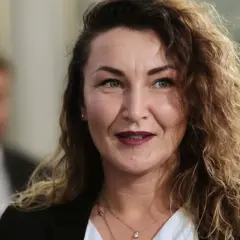 Monika Pawłowska z kręconymi włosami, w białej bluzce i czarnej marynarce, uśmiecha się.