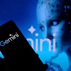 Ręka trzyma smartfon z logo Gemini. W tle widać twarz robota, która przypomina miękkie dildo.
