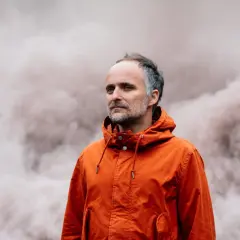 Artur Rojek w pomarańczowej kurtce, na tle dymu. Jego twórczość często porusza tematykę przemijania i refleksji.