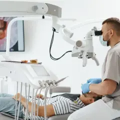 Stomatolog w masce i rękawiczkach bada pacjenta pod mikroskopem. Na ekranie widać zbliżenie zębów.
