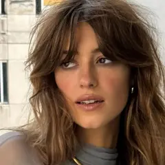 Mujer con flequillo cortina desfilado, cabello castaño ondulado y mirada pensativa.