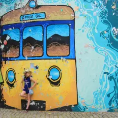 Dziewczynka w kapeluszu pozuje przy kolorowym muralu z tramwajem, który jest jedną z opolskich atrakcji dla dzieci.