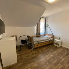 Pokój w domu seniora z łóżkiem, chodzikiem, szafką i telewizorem.