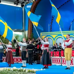 Människor i folkdräkter viftar svenska flaggor under firandet av 6 juni i Stockholm. En orkester spelar i bakgrunden.