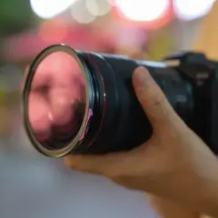 Dłoń trzyma aparat z obiektywem, gotowy do uchwycenia chwili. Czerwony pierścień obiektywu i fioletowy element przy **shutterze** dodają mu charakteru.
