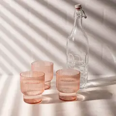 Natural product photography: three pink ribbed glasses filled with water and a clear glass bottle with a swing top, bathed in dappled light.