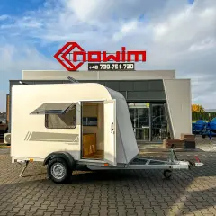 Mały, biały kamper z przyczepą, z otwartymi drzwiami i oknem, stoi przed budynkiem z czerwonym logo.