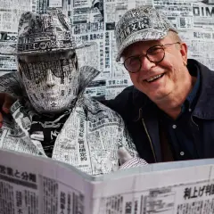 Tomaszewski fotograf uwiecznił artystę ubranego w gazety. Obok uśmiechnięty mężczyzna w okularach.