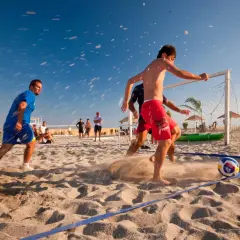 Mężczyźni grają w piłkę nożną na plaży, pokazując różne odmiany tej gry. Słońce, piasek i błękitne niebo tworzą idealne warunki do sportowych emocji.