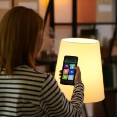 Kobieta steruje domem za pomocą smart life app na telefonie. Widok zza ramienia na ekran z ikonami.