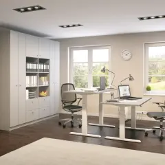 Modernes Arbeitszimmer einrichten: Zwei höhenverstellbare Schreibtische mit Laptops, Bürostühle, ein großer Schrank und Fenster mit Blick ins Grüne.