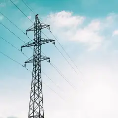 Wysoki słup energetyczny z przewodami, symbolizujący transport przesyłowy energii elektrycznej na tle błękitnego nieba z chmurami.