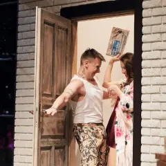 Scena ze spektaklu "Pomoc domowa" w Radomiu. Mężczyzna w podkoszulku i kolorowych spodniach trzyma gazetę, kobieta w kwiecistej sukience śmieje się.