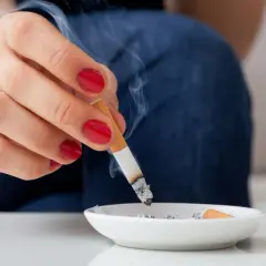 Eine Hand mit roten Nägeln hält eine brennende Zigarette über einem Aschenbecher. Rauch steigt auf und hinterlässt einen kalter rauch geruch.