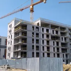 Budowa nowych mieszkań na sprzedaż w Bochni. Dwa bloki w budowie, jeden prawie gotowy, drugi w stanie surowym. Dźwigi budowlane na tle błękitnego nieba.