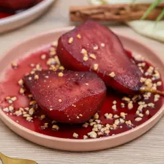 Peras al vino, tiernas y brillantes, servidas en un plato rosa con trocitos de nuez y un sirope rojo intenso.