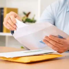 Dłoń wkłada białą kartkę do koperty. Czy można wysłać list bez nadawcy? Na biurku leżą koperty.